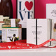 A close-up view of a gift arrangement featuring a jar of salted nut mix, a candle labeled "Lemongrass & Ginger," and decorative red ribbon that reads "All my love." In the background, there are bottles and other gifts with a romantic theme. A close-up view of a gift arrangement featuring a jar of salted nut mix, a candle labeled "Lemongrass & Ginger," and decorative red ribbon that reads "All my love." In the background, there are bottles and other gifts with a romantic theme.